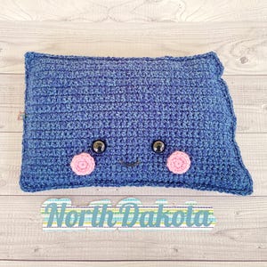 May include: A blue crocheted cushion shaped like the state of North Dakota with a smiling face, pink cheeks, and black eyes. The cushion has the text "North Dakota" written below it.