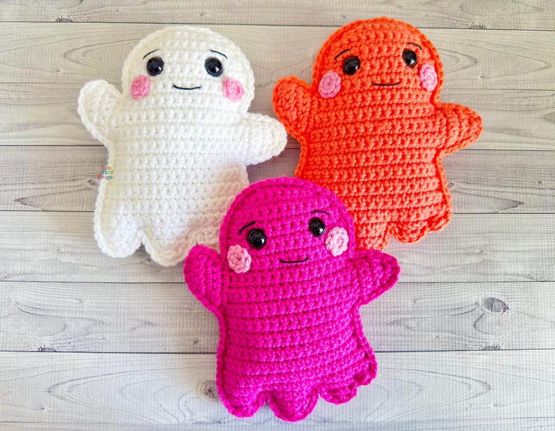 May include: Three crocheted ghost plushies. One is white, one is orange, and one is pink. Each ghost has a smiling face with black eyes and pink cheeks.