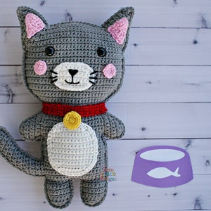May include: A gray crocheted cat toy with pink ears, a red collar, and a yellow bell. The cat is sitting on a white wooden surface with a purple food bowl with a white fish inside.