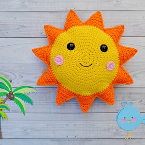 Puede incluir: Una almohada de sol de crochet con una cara sonriente, rayos naranjas y mejillas rosadas. El sol está sobre un fondo de madera blanca con dos palmeras de papel y un pájaro azul de papel.
