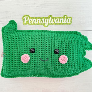 May include: Green crocheted pillow shaped like the state of Pennsylvania with a smiling face, black eyes, and pink cheeks. The pillow has the word "Pennsylvania" written on it.