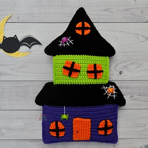 May include: A crocheted Halloween house cushion with a black roof, green middle section, and purple bottom. The house has orange windows and a door, and there are spiders and spiderwebs on the roof and sides.