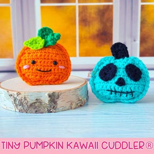 May include: Two crocheted pumpkin-shaped toys. One is orange with a green stem and smiling face. The other is turquoise with black eyes, nose, and mouth. The text "TINY PUMPKIN KAWAII CUDDLER" is at the bottom.