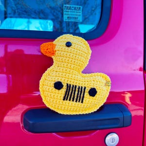 Peut inclure: Un canard en caoutchouc jaune crochet&eacute; avec des yeux noirs et un motif de calandre Jeep noir et blanc sur la poitrine. Le canard est attach&eacute; &agrave; la poign&eacute;e d'une porti&egrave;re de voiture rouge.