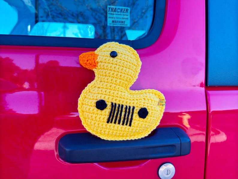 Peut inclure: Un canard en caoutchouc jaune crochet&eacute; avec des yeux noirs et un motif de calandre Jeep noir et blanc sur la poitrine. Le canard est attach&eacute; &agrave; la poign&eacute;e d'une porti&egrave;re de voiture rouge.