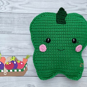 May include: A green crocheted plush bell pepper with a smiling face, black eyes, and pink cheeks. The pepper has a green stem on top.