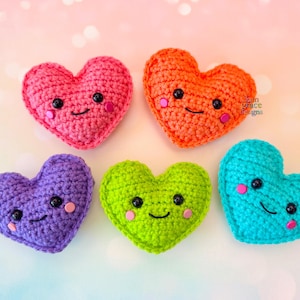 Puede incluir: Cinco corazones de ganchillo coloridos con caras sonrientes. Los corazones son en tonos de rosa, naranja, morado, verde y turquesa. Cada corazón tiene ojos de botón negros y mejillas rosadas. Los corazones están dispuestos sobre un fondo suave y borroso.