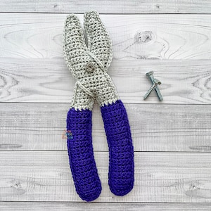 May include: A handmade crochet pair of pliers in grey and blue, with a silver button detail. The handles are a deep blue. Two silver screws are next to the pliers. The item is likely a toy or decorative piece.