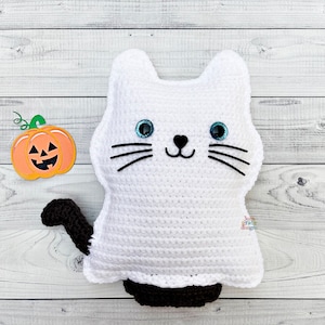 Op de afbeelding: Een witte gehaakte spookkat met zwarte ogen en een zwarte neus. De kat heeft een zwarte staart en houdt een kleine oranje pompoen vast met een pompoengezicht.