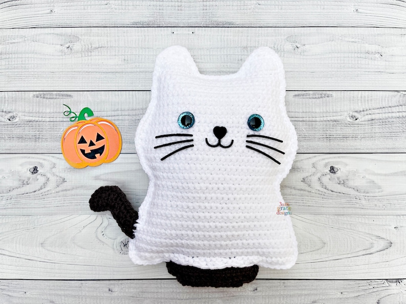 May include: A white crocheted ghost cat with black eyes and a black nose. The cat has a black tail and is sitting on a white wooden surface. A small orange pumpkin with a green stem is to the left of the cat.