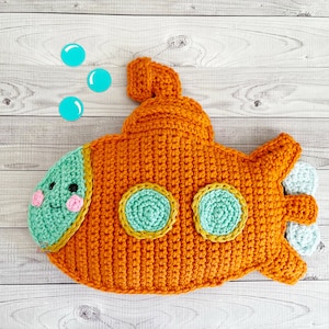 May include: A handmade orange crochet submarine with turquoise accents, including portholes and a smiling face on the front. The submarine has light blue fins and is set against a light wood background with three turquoise bubbles.