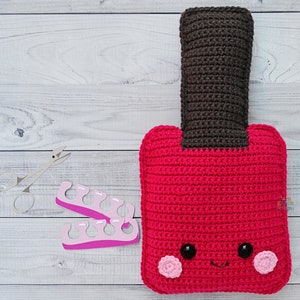 May include: A crocheted red nail polish bottle with a black cap and a smiling face. The bottle has two pink cheeks and black eyes. There are also two pink nail separators and a pair of nail clippers on a white wooden surface.