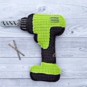 Może przedstawiać: Zabawka w kształcie wiertarki wykonana na szydełku w kolorze zielonym i czarnym z szarym wiertłem. Wiertarka leży na białym drewnianym podłożu, obok niej znajdują się dwa małe wiertła.