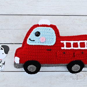 May include: A red crocheted fire truck toy with a blue windshield, white ladder, and black wheels. The truck has a smiling face with a black eye, a pink cheek, and a white light on top.
