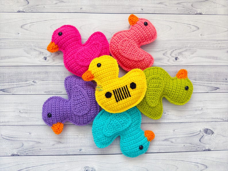 Peut inclure: Un groupe de six jouets en crochet en forme de canard de diff&eacute;rentes couleurs, dont le rose, le violet, le jaune, le vert et le bleu. Les canards sont dispos&eacute;s en cercle sur une surface en bois blanc.