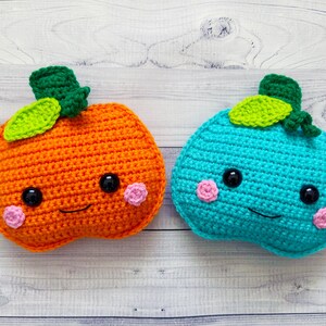 Op de afbeelding: Twee gehaakte pompoenen met lachende gezichten. De ene pompoen is oranje met groene bladeren en de andere is turquoise met groene bladeren.