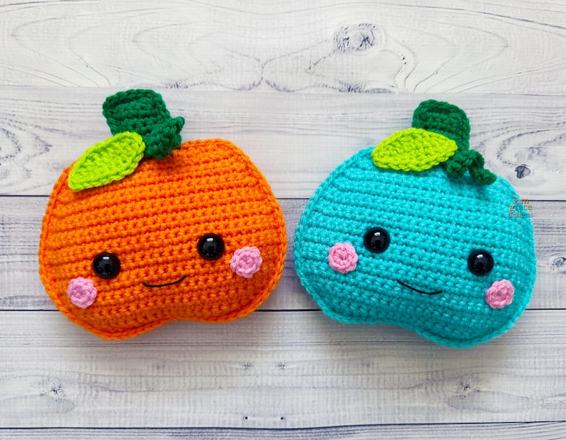 Op de afbeelding: Twee gehaakte pompoenen met lachende gezichten. De ene pompoen is oranje met groene bladeren en de andere is turquoise met groene bladeren.