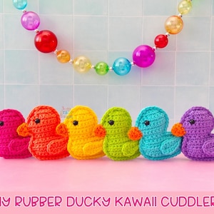 Puede incluir: Seis patitos de ganchillo en fila, cada uno de un color diferente: rosa, naranja, amarillo, verde, turquesa y morado. Tienen picos naranjas y ojos negros. Una colorida cadena de cuentas cuelga encima. El texto "TINY RUBBER DUCKY KAWAI CUDDLER" está en la parte inferior.