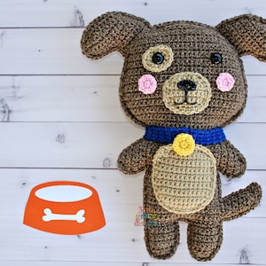 May include: A brown crocheted dog toy with pink cheeks, black eyes, and a blue collar with a yellow button. The dog is standing on a white wooden surface with an orange dog bowl with a white bone.