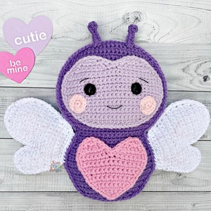 Puede incluir: Una abeja de crochet morado con alas blancas y un corazón rosa en el pecho. La abeja tiene una cara sonriente con ojos negros y mejillas rosas. El texto "be mine" y "cutie" está escrito en dos corazones rosas.