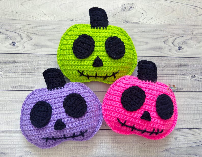 Op de afbeelding: Drie gehaakte Halloween-pompoenen met zwarte ogen en gestikte monden. E&eacute;n pompoen is groen, &eacute;&eacute;n is paars en &eacute;&eacute;n is roze.