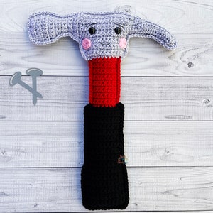 May include: A crocheted hammer toy with a red and black handle and a smiling face. The hammer is on a white wooden background with two small crocheted nails.