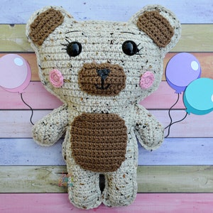 May include: A crocheted brown and white teddy bear holding pink, purple, and blue balloons. The bear has brown eyes, a brown nose, and pink cheeks.