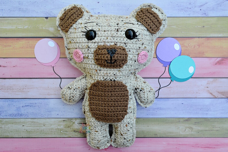 May include: A crocheted brown and white teddy bear holding pink, purple, and blue balloons. The bear has brown eyes, a brown nose, and pink cheeks.