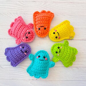 May include: A collection of seven colorful, crocheted creatures with smiling faces. The creatures are in shades of pink, orange, yellow, purple, teal, and green. Each has black button eyes and a stitched smile. The items are arranged in a circle.