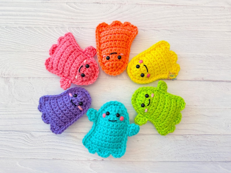 May include: A collection of seven colorful, crocheted creatures with smiling faces. The creatures are in shades of pink, orange, yellow, purple, teal, and green. Each has black button eyes and a stitched smile. The items are arranged in a circle.