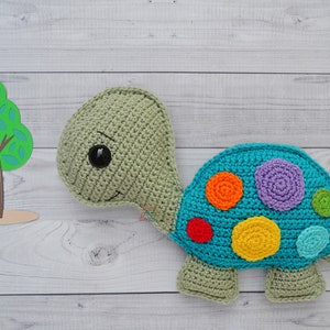Puede incluir: Una tortuga de ganchillo hecha a mano en tonos verdes y turquesa, con un caparazón colorido adornado con aplicaciones en forma de flor naranja, morada, roja y amarilla. La tortuga tiene una cara sonriente y un ojo negro grande.