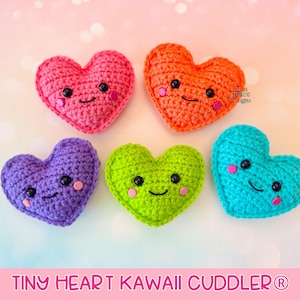 Może przedstawiać: Pięć szydełkowych zabawek w kształcie serca w różnych kolorach: różowym, pomarańczowym, fioletowym, zielonym i niebieskim. Każde serce ma uśmiechniętą twarz z czarnymi oczami i różowymi policzkami. Tekst "TINY HEART KAWAII CUDDLER®" znajduje się na dole.