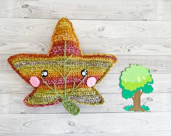 May include: A crocheted leaf with a smiling face, pink cheeks, and a green stem. The leaf is multi-coloured with shades of orange, red, and green. The leaf is sitting on a white wooden surface next to a green paper cutout of a tree.
