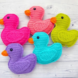 Peut inclure: Cinq canards en crochet de diff&eacute;rentes couleurs&nbsp;: rose, vert, violet, bleu et orange. Les canards sont dispos&eacute;s en cercle sur un fond en bois blanc.