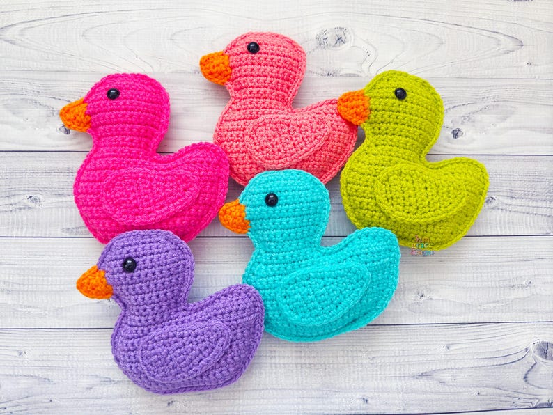 Peut inclure: Cinq canards en crochet de diff&eacute;rentes couleurs&nbsp;: rose, vert, violet, bleu et orange. Les canards sont dispos&eacute;s en cercle sur un fond en bois blanc.