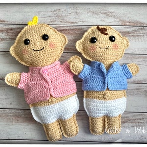 May include: Two crocheted baby dolls, one wearing a pink cardigan and the other wearing a blue cardigan. Both dolls are wearing white crocheted diapers. The dolls are sitting on a wooden surface.