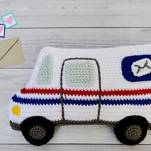 Peut inclure: Un coussin en forme de camion postal crocheté blanc avec des rayures rouges, bleues et blanches, une fenêtre verte, une porte grise et un logo postal bleu avec une lettre "M" grise.