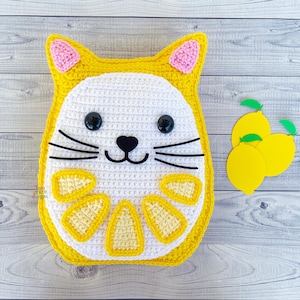 May include: A crocheted cat-shaped pillow with a lemon design. The pillow is yellow and white with black eyes and whiskers.