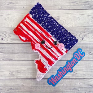 May include: A crocheted plush toy shaped like the outline of Washington, D.C. The toy is decorated with red, white, and blue yarn to resemble the American flag. The toy has a smiling face with black eyes and pink cheeks. The toy is sitting on a wooden surface with a blue sign that says "Washington DC".