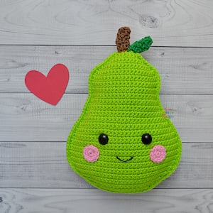 Op de afbeelding: Een handgemaakt, groen gehaakt peer-vormig kussen met een lachend gezicht, zwarte ogen en roze wangen. De peer heeft een bruine steel en groene bladeren. Een rood hart ligt naast de peer op een witte houten achtergrond.