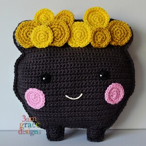 May include: A black crocheted pot with a smiling face and pink cheeks. The pot is topped with a crown of yellow crocheted coins.