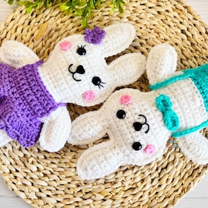 May include: Two crocheted bunny plushies, one wearing a purple dress and the other wearing a turquoise outfit. The bunnies are sitting on a woven straw mat.