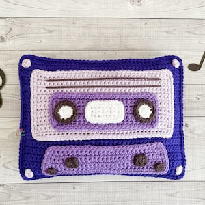 May include: A purple and white crocheted cushion shaped like a cassette tape. The cushion has a brown crocheted tape and a white crocheted label.