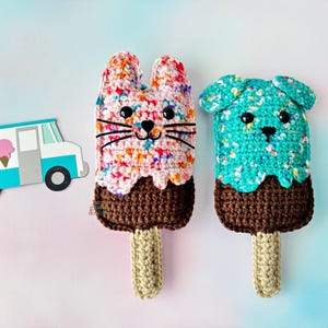 May include: Two handmade crochet ice pop toys, one shaped like a bunny and the other a dog, with brown "chocolate" bottoms and beige sticks. The bunny is multicolored, and the dog is turquoise. A paper ice cream truck is on the left.