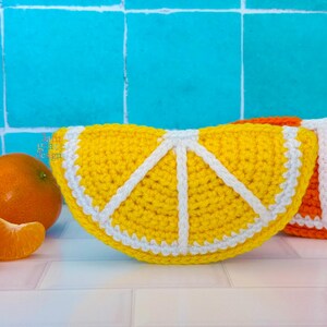 May include: A yellow crochet citrus slice with white detailing and a white border, positioned next to a partial orange crochet slice and fresh mandarin oranges. The yellow slice features a segmented design with white yarn lines.