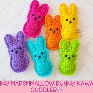 Puede incluir: Seis conejitos de ganchillo de colores brillantes: rosa, naranja, amarillo, morado, turquesa y verde. Cada conejo tiene ojos de cuentas negras. El texto "TINY MARSHMALLOW BUNNY KAWAII CUDDLER" aparece en la parte inferior.