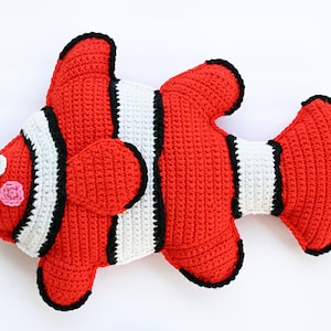 May include: A crocheted stuffed clownfish toy with red and white stripes and black accents. The fish has a pink cheek and a black eye.