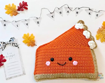 May include: A crocheted pumpkin pie slice pillow with a smiling face, white whipped cream, and a brown crust. The pillow is surrounded by fall leaves and a banner that reads "GIVE THANKS".