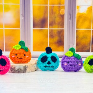 May include: Five crocheted pumpkins in pink, orange, teal, purple, and green. Each pumpkin has a black face and a green stem. The pumpkins are displayed on a wooden slice in front of a window with a blurred background.