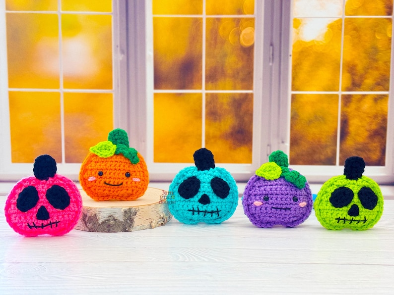May include: Five crocheted pumpkins in pink, orange, teal, purple, and green. Each pumpkin has a black face and a green stem. The pumpkins are displayed on a wooden slice in front of a window with a blurred background.